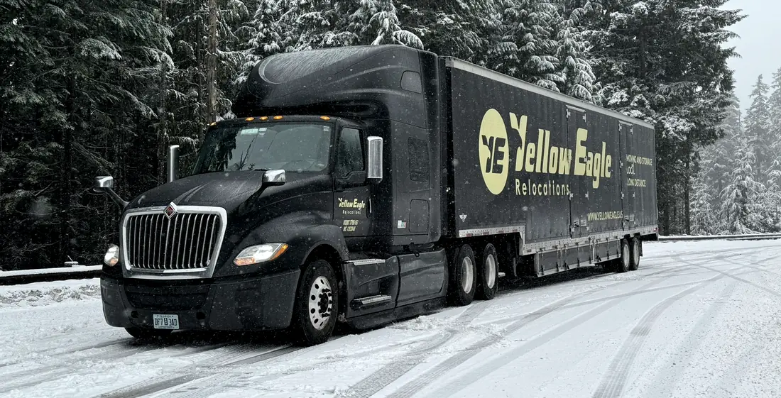 A Yellow Eagle tractor and trailer on a snow-covered road between snow-laden evergreens