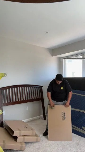 A mover in a Yellow Eagle shirt closing a carton marked GLASS in a bedroom